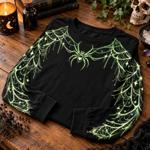 Folded black Glow Spider Web Long Sleeve tee on wooden table with pale green spider and web artwork visible